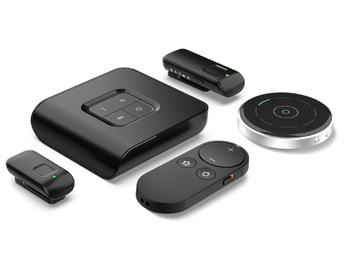 A collection of sleek, modern hearing aid accessories and assistive listening devices displayed on a white background. The set includes a central square unit with control buttons, a circular remote microphone, and various small wireless devices, all in black or silver with minimalist designs and LED indicators, suggesting advanced connectivity and user-friendly operation.
